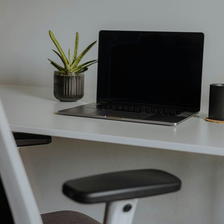 Aufgeräumter weißer Schreibtisch mit geöffnetem Laptop, Pflanze und Schreibtischstuhl